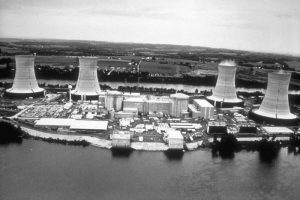 a black and white photograph of Three Mile Island showing four large cooling towers in the background and the Susquehanna river in the foreground.