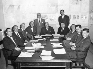 black and white photograph of eleven men in suits, nine sitting and two standing, around a conference table