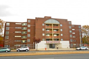 A color photograph of the Guild House building. The image shows the brick building and the sidewalk and street in front of the building. There are three cars parked in front of the building. 