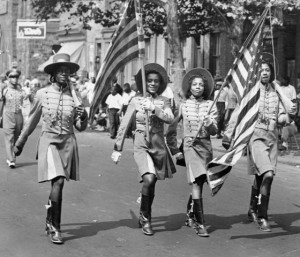 A black and white photograph depicting four African American women walking in color guard garb displaying American flags. They are walking as part of a larger parade, observers are lining the sidewalk in the background.