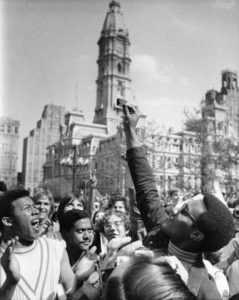 A young African American man wearing glasses, a turtleneck and jacket holds his burning draft card up above a crowd of onlookers. The draft card is held in front of City Hall (as it is framed), making a symbolic image. (City Hall and other buildings can be seen in the background.) Another African American man to the left claps with a look of satisfaction on his face as he looks upward at (likely) his friend's burning draft card.