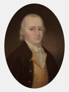 White man with blonde hair who's balding on the sides of head. He is wearing a brown coat and yellow waistcoat on top of a white, ruffled undergarment.