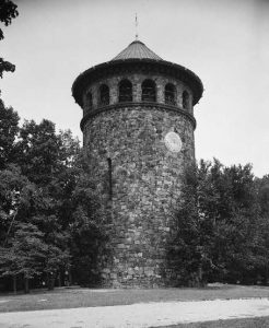 A black and white photograph of Rockford tower, a tall cylindrical stone tower with a pyramidal roof. The upper floors are lined with arched windows. There is a sundial embedded in the stone below the windows. The tower stands in a park surrounded on two sides by trees, with a walking path leading towards it.