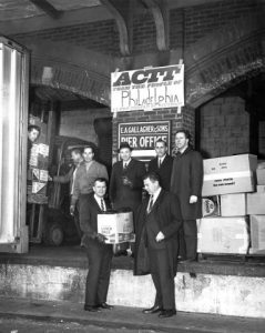 A group of well-dressed men stand in front of boxes of supplies to be donated to South Vietnam. The boxes have a variety of markings, although most indicate canned goods such as soup (although one is marked with Schmidt's of Philadelphia Brewery). A sign behind them indicates the Pier Office, and a sign above that reads "ACTT: FROM THE PEOPLE OF PHILADELPHIA TO THE PEOPLE OF VIETNAM." A few men help load a truck just out of frame to the left.