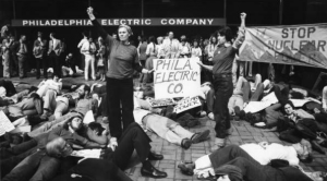 a black and white photograph of a group of protesters in front of the PECO building. Most of the protesters lay on the ground as if dead. Two in the front hold a sign reading "Phila. Electric Co." with their fists raised in the air. Behind them, a large banner reads "Stop nuclear power"