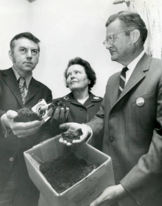 a black and white photograph of Ruth Patrick flanked by two unidentified men. The man on the right is holding a cardboard box of soil. All three people are holding soil samples in their outstretched hands and are wearing buttons with Earth Day logos on them.