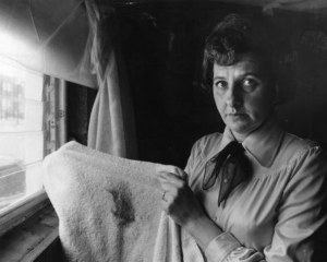 a black and white photograph of a woman standing in front of a window. She is looking direcltly into the camera and holding up a white towel with a large black soot mark on it.